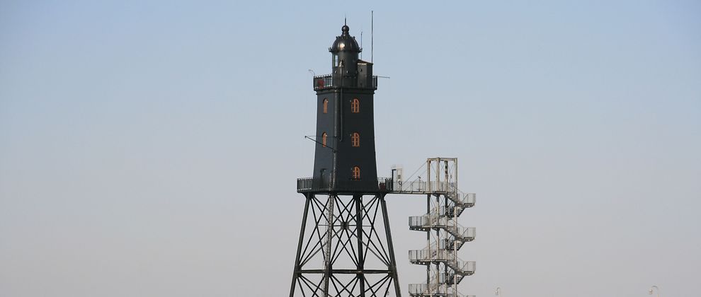  Ein Leuchtturm steht auf Stelzen im Wasser. Er ist schwarz und außen führt eine Treppe auf den Leuchtturm hinauf.