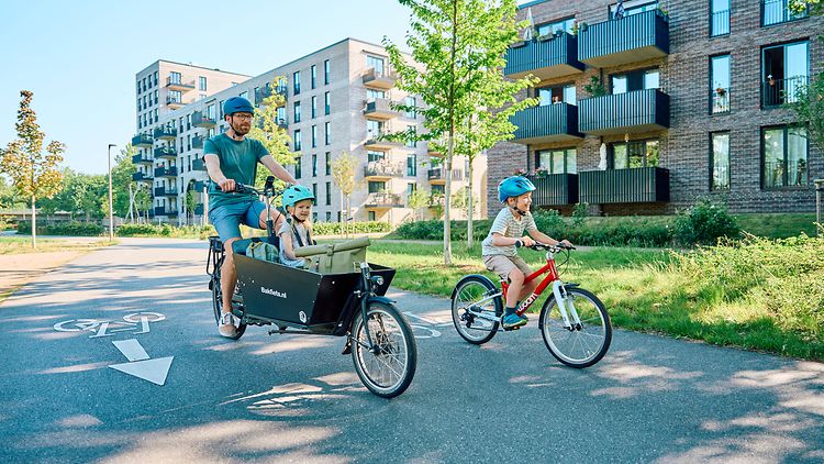 Mann mit Lastenrad unterwegs mit zwei Kindern auf einer breiten Fahrradstraße im Wohngebiet