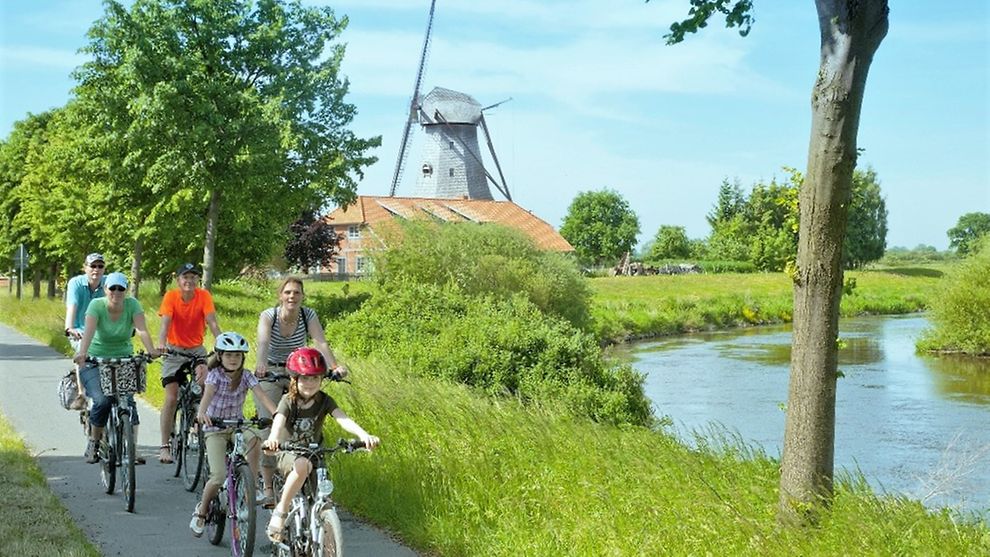  Radfahrende Familie am Fluss mit Windmühle im Hintergrund