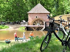  Radfahrer und Wasserwanderer am Fluss mit Wassermühle im Hintergrund