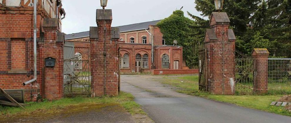  Auf einem Fabrikgelände sind große Gebäude aus rotem Backstein zu sehen. Im Vordergrund des Bildes ist ein Eingangsbereich zum Gelände zu sehen. Es ist ein geöffnetes Tor.
