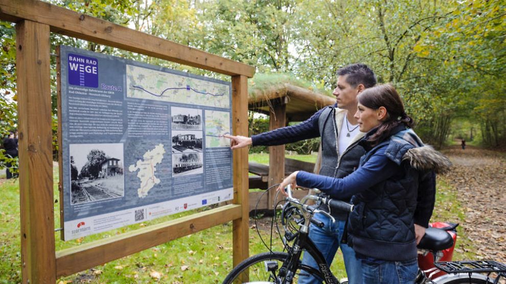Two cyclists standing in front of an information point along a path in the woods.