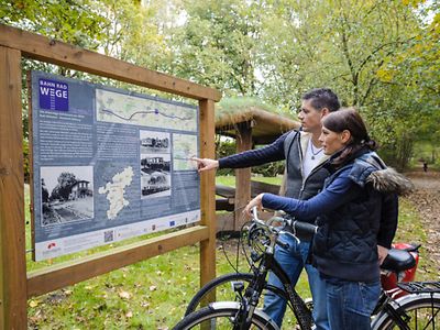  Two cyclists standing in front of an information point along a path in the woods.