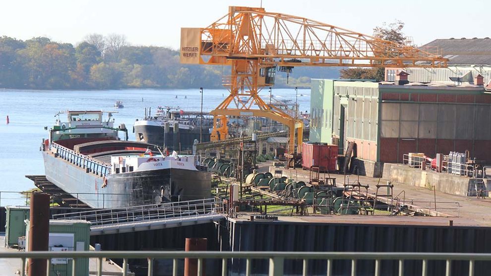 Ein Schiff liegt am Gelände der Hitzler Werft. Es ist von Wasser umgeben. Zu sehen sind Maschinen und ein gelber Kran.