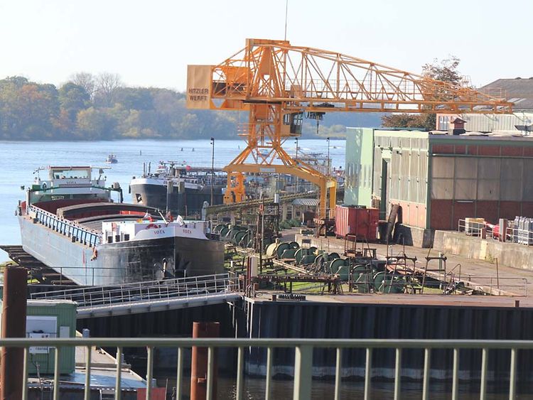  Ein Schiff liegt am Gelände der Hitzler Werft. Es ist von Wasser umgeben. Zu sehen sind Maschinen und ein gelber Kran.