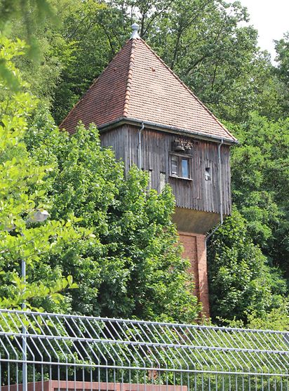 Ein Turm, dessen oberer Teil mit Holz verkleidet ist, steht hinter einem Zaun umgeben von Bäumen.