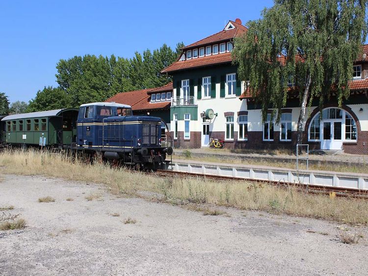  Ein Zug mit einer blauen Lok steht vor einem weiß-roten Gebäude. Um den Zug herum wachsen Gräser.