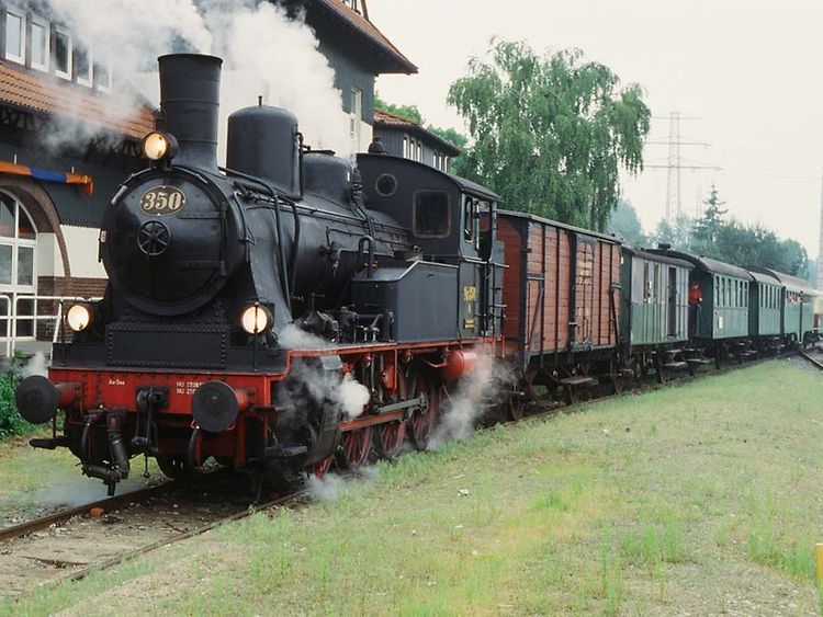  Ein Zug mit einer schwarz-roten Lok und mehreren Waggons in grün und rot steht auf einem Gleis. Weißer Dampf kommt aus der Lok. Es ist die Museumsbahn Karoline.