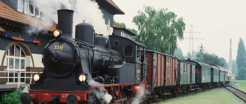  Ein Zug mit einer schwarz-roten Lok und mehreren Waggons in grün und rot steht auf einem Gleis. Weißer Dampf kommt aus der Lok. Es ist die Museumsbahn Karoline.