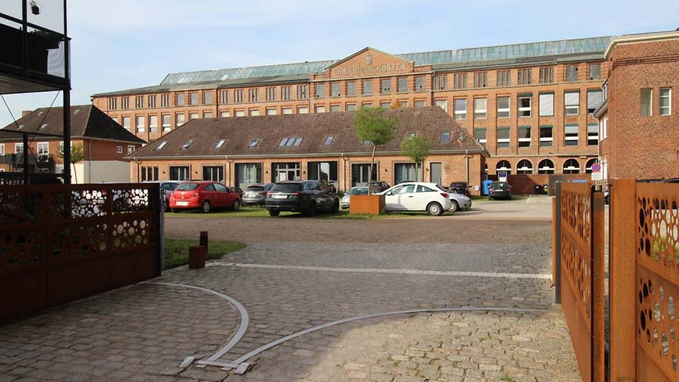 Die ehemalige Tuchfabrik Christian Friedrich Köster ist ein mehrstöckiges großes rotes Gebäude mit vielen Fenstern. Davor ist ein Parkplatz und ein kleineres Gebäude.