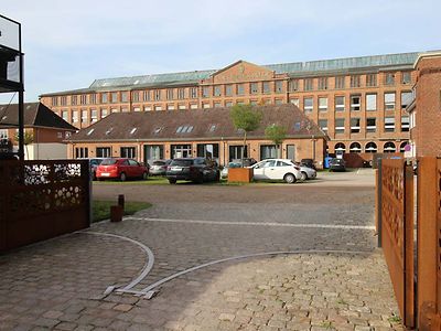  Die ehemalige Tuchfabrik Christian Friedrich Köster ist ein mehrstöckiges großes rotes Gebäude mit vielen Fenstern. Davor ist ein Parkplatz und ein kleineres Gebäude.