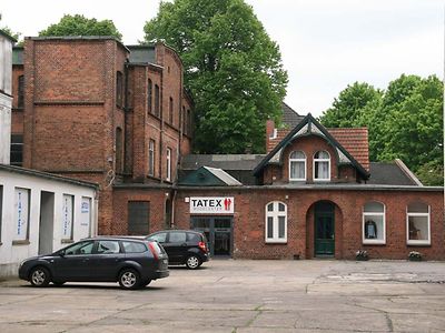  Ein roter Backsteinbau ist die ehemalige Tuchfabrik Julius Bartram. Der Eingangsbereich ist ein kleineres Gebäude, links davon befindet sich ein mehrstöckiger Gebäudeteil.