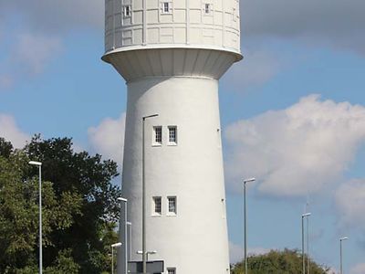  Der Wasserturm ist ein weißer Turm, der oben breiter ist als unten. Er steht hinter einer Mauer.