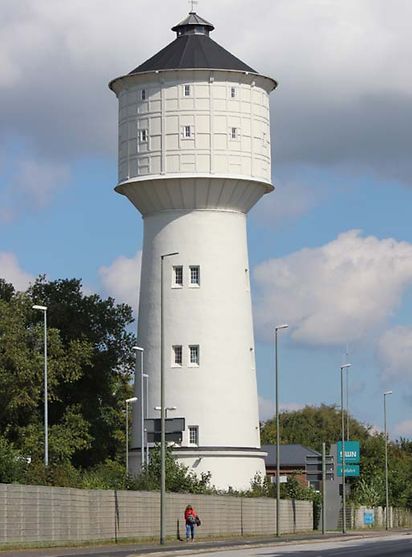Der Wasserturm ist ein weißer Turm, der oben breiter ist als unten. Er steht hinter einer Mauer.