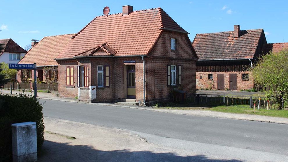Ein kleines rotes Haus mit einem vorbei und einem rot gedeckten Dach steht an einer Straße. Es ist das Chausseeeinnehmerhaus Mueß.