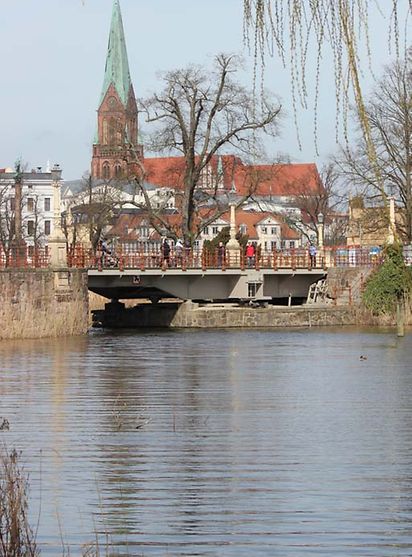 Eine Brücke führt über Wasser. Im Hintergrund sind Häuser und eine Kirche zu sehen.