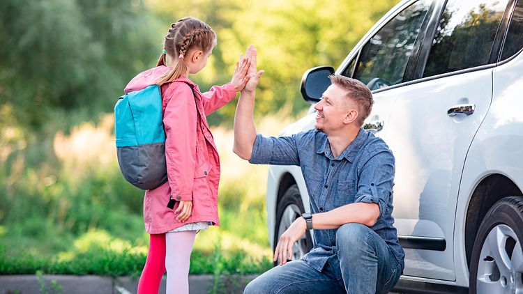  Ein Mann und ein Mädchen verabschieden sich auf dem Weg zur Schule, im Hintergrund eine Auto